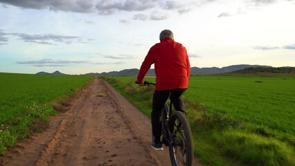 Elderly man on an electric mountain bike doing sports through a green field. He is on a path and in the background are some mountains. Cloudy day at sunset. Concept seniors in active, enjoying life