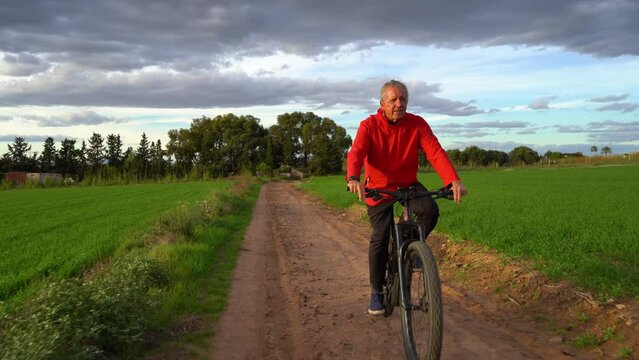 Senior man on electric mountain bike doing sports through a green field. He is on a road and in the background a wooded area and clouds. Cloudy day at sunset. Concept seniors in active, enjoying life