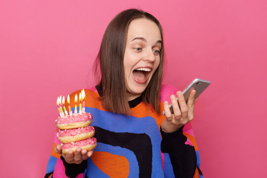 Image of excited woman holds tasty doughnuts with burning candles and smart phone, reading message with congratulations, wears sweater, celebrating birthday alone, standing isolated on rose background