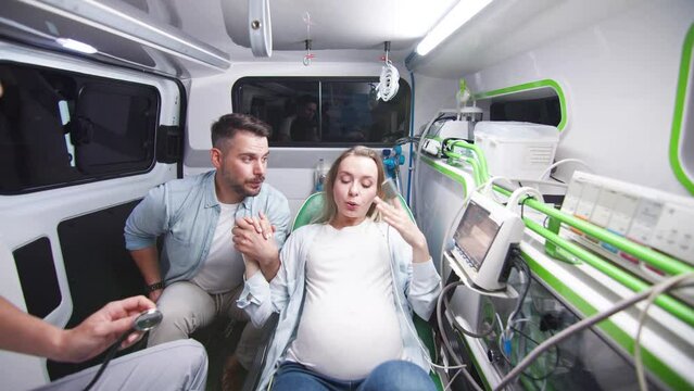 Young pregnant woman having labor contractions lying on stretchers in ambulance with female paramedic providing help and husband. Witty man trying to do selfie holding hand of wife supporting.