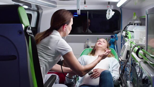 Professional multiethnic paramedics providing help measuring heartbeat of baby with stethoscope pregnant woman during delivery. Caucasian female patient shouting while labor contractions in ambulance.
