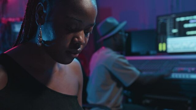 Tilt up of young black woman in smart casualwear pressing keys of electric synthesizer while recording new music with sound engineer sitting by mixing board and adjusring noise level