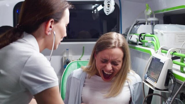Close-up of female paramedic providing help for young woman with labor contractions in ambulance. Young female patient suffering and shouting. Nurse listening to heartbeat of baby with stethoscope.