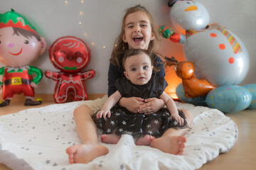 Two cute little sisters sitting on the play mat with funny expression. Christmas decoration in the...