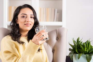 Healthy lifestyle concept, caucasian beautiful millennial woman healthy lifestyle concept. Long brown hair girl sitting on armchair at home holding glass of fresh clean water. Looking camera smiling.