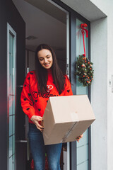 Vertical photo - Smiling woman in Christmas sweater holding a box at the front door waiting for the mailman to come