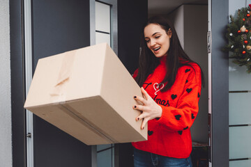 Smiling woman in Christmas sweater exited about the package she is holding in her hands while standing at the front door