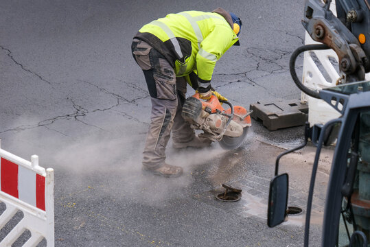 Ein Arbeiter in Sicherheitskleidung schneidet auf einer abgesperrten Baustelle die Asphaltdecke einer Stra&szlig;e mit einer Kreiss&auml;ge auf, Luftaufnahme, Bagger, Maschinen und Werkzeuge