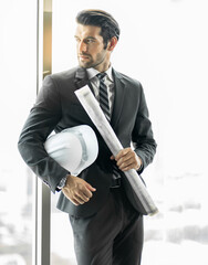 Portrait of handsome engineer manager in dark suit holding construction blueprint and white helmet...