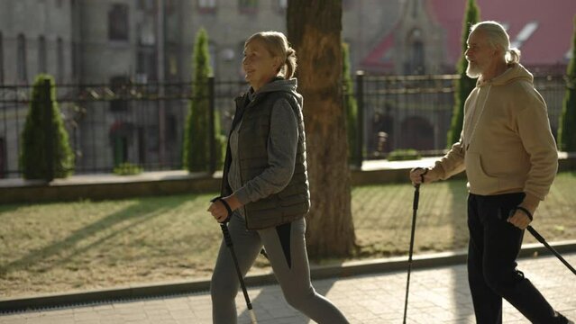 Happy senior couple with trekking poles walking in the street. Practicing together Nordic walking outdoors. Aging youthfully. Elderly people and sport concept.