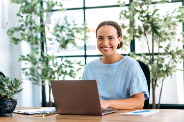 Happy lovely mixed race business lady, sitting at a desk in the modern office, working in a laptop on a project, browsing the information on the Internet, looking at camera, smiling friendly