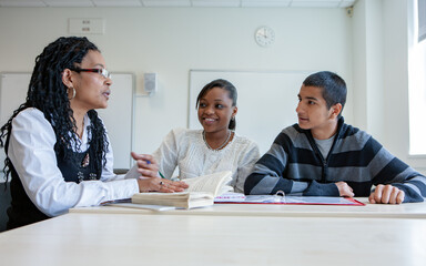 College Class: Listening to Teacher. A pair of late teenage students working with their teacher in the classroom. From a series of related images.