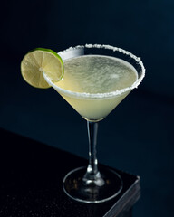 Cocktail Margarita. Alcoholic drink with tequila, lime juice and citrus liqueur. Drink in cocktail glass, decorated with lime on table. Dark background. Copy space. 