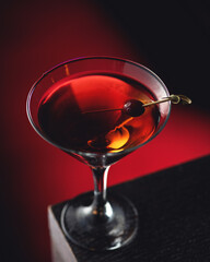 Alcoholic cocktail Manhattan with whiskey and vermouth served with cherry. Alcoholic drink in cocktail glass on edge of table. Dark red backlit background. Soft focus. View from above. Close-up.