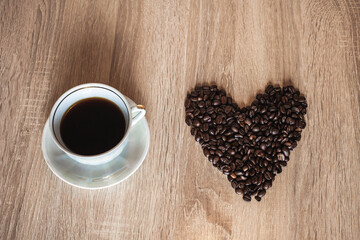 A cup of hot aromatic coffee in a saucer, against the background of scattered coffee beans in the form of a heart, on a wooden light brown table.