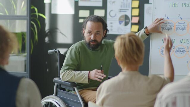 Male corporate trainer in wheelchair explaining presentation on flipchart to office employees while providing business education