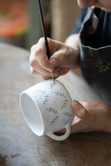 Hands of professional artisan hold white ceramic mug drawing pattern with dark paint using brush on blurred background. Woman enjoys working with handmade pottery craft in studio extreme closeup - obrazy, fototapety, plakaty