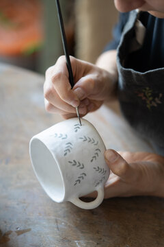 Hands of professional artisan hold white ceramic mug drawing pattern with dark paint using brush on blurred background. Woman enjoys working with handmade pottery craft in studio extreme closeup