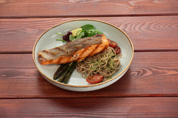 Grilled salmon steak served with grilled tomatoes, salad, lemon sliced, pepper seeds, rosemary, and parsley on a plate on a wooden table.