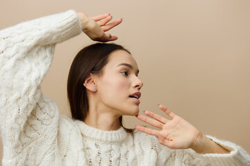 an elegant woman stands on a light background in a white sweater gently raises her hand above her head, placing it on her hair, brings the second one to her cheek and gently touches it