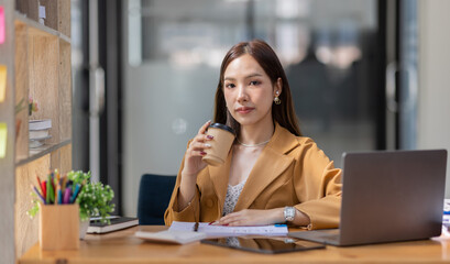 Happy Asian young woman drinking coffee while working online in office, employee freelance online marketing e-commerce telemarketing concept.
