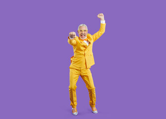 Funny energetic senior man dancing and having fun in studio. Happy cheerful old man wearing bright yellow suit enjoying music and dancing isolated on solid purple background. Party and fashion concept