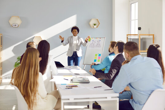 Business Team Having A Meeting, Sitting Together At White Office Table And Listening To Black Woman Standing At Whiteboard Making Presentation On Soft Skills, Management, And Communication Strategies