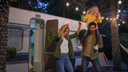 Caucasian funny family in camping in forest near van automobile trailer dancing in evening night near lights. Man father holding kid daughter on neck shoulders dace with wife woman mother celebrate