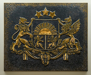 Closeup of gold color coat of arms of Latvia embossed on a relief dark metal plate.