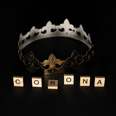 Golden crown on black background, in the foreground written in wooden letters CORONA, COVID-19 mood picture.