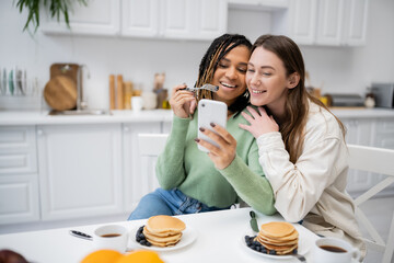 cheerful african american lesbian woman showing smartphone to happy girlfriend during breakfast.