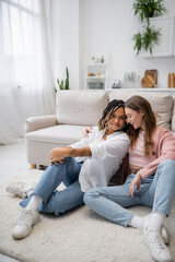 young lesbian woman sitting with tattooed african american girlfriend on carpet near couch.