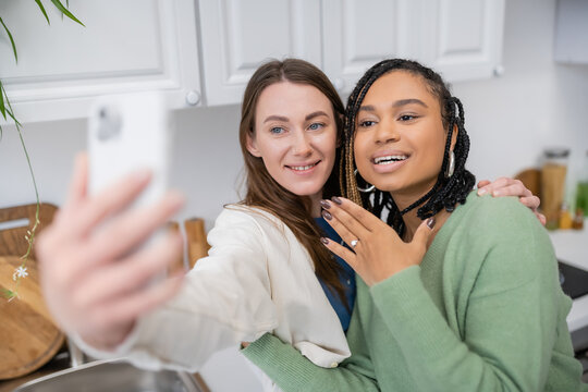 lesbian woman taking selfie while cheerful african american girlfriend showing engagement ring on finger.
