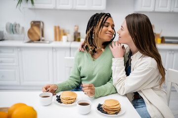 cheerful lesbian woman hugging african american girlfriend during breakfast.