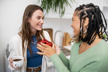 african american lesbian woman holding red gift box near happy girlfriend with cup of coffee on valentines day.