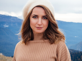 beautiful blonde in a beige sweater and beret looks at the camera and smiles. Portrait of a cute woman in the mountains