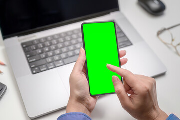 woman using smartphone with green screen over laptop in office.