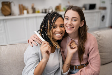 happy african american and lesbian woman holding pregnancy test near smiling girlfriend.