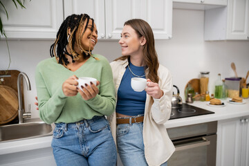 cheerful multiethnic lesbian couple holding cups of coffee and looking at each other in kitchen.