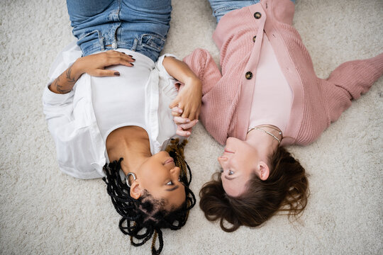 top view of positive and multiethnic lesbian couple lying on soft carpet and holding hands.