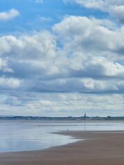 The houses, buildings and church steeple of a small coastal town form a thin ribbon dividing a huge, clouded sky from its reflection in sea and sand.