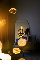 Merry Christmas and Happy Holidays! Young african american woman sitting in an armchair and drinking coffee by the Christmas tree in a cozy living room at home