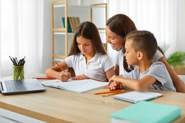 Smiling supported mom helping her children doing homework sitting at desk. Girl is writing in copybook and boy is looking at her and smiling. Homeschooling, family education, online studying concept.