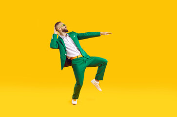 Fototapeta na wymiar Cool and stylish young man is having fun dancing celebrating Saint Patricks Day. Caucasian man in stylish green suit, white shirt and sunglasses smiling while dancing on orange background. Web banner