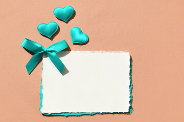Valentine's Day background. A blank white piece of paper with a turquoise bow and decorative hearts on a beige background. Flat layout, space for text.