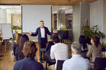 Business trainer in suit and shirt is greeting group of adult business people students in modern office spreading his arms asides. People sitting at desks backs to camera. Adult education concept.