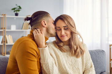 Portrait of handsome husband who gently kissing his attractive wife in cheek at home in morning. Couple is celebrating Valentine's Day. Concept of human emotions, love, relations and romantic holidays