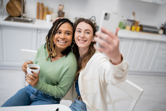 cheerful lesbian woman taking selfie on smartphone with joyful african american girlfriend.
