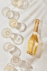 Flat lay with white sparkling wine bottle, set glasses wine with sunshine shadow palm leaf on light...