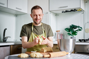 Fototapeta na wymiar Attractive caucasian bearded chef baker in green apron working with dough on wooden cutting board with rolling pin on foreground. Working at home kitchen concept, homemade baking. High quality photo